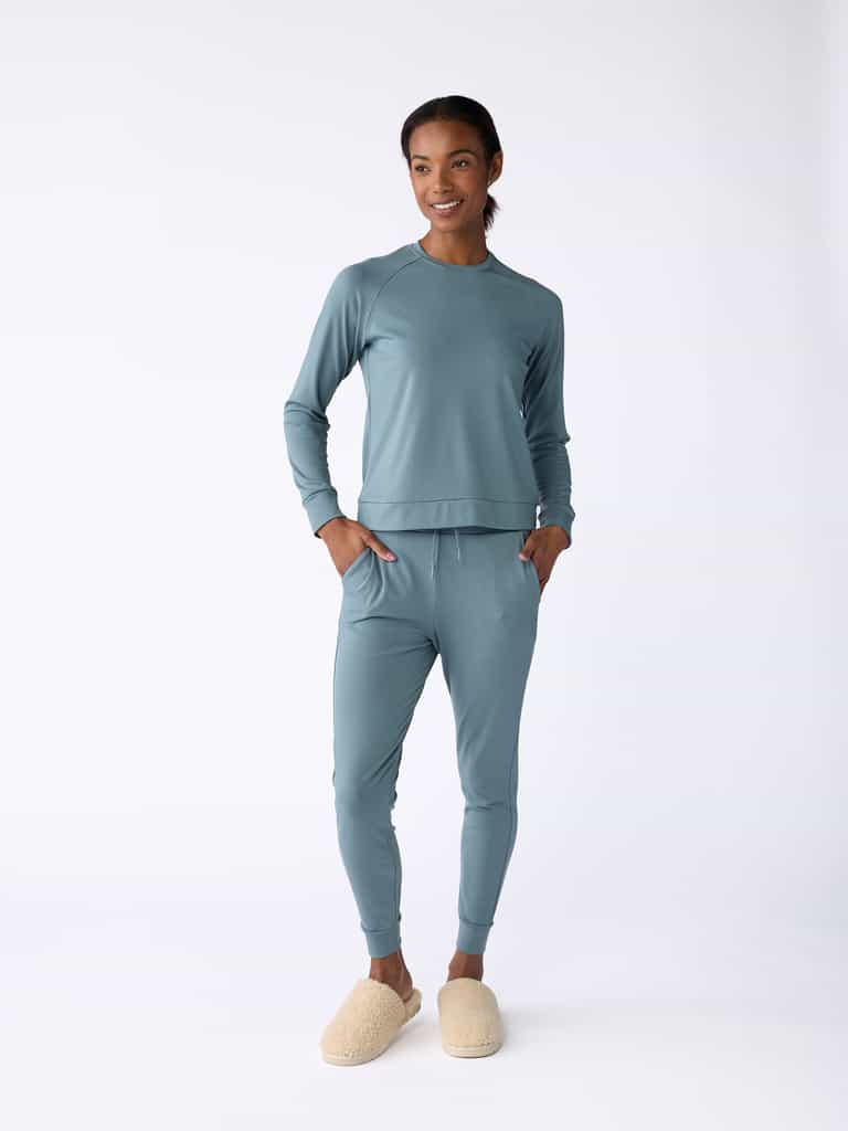 A person in a matching light blue ensemble, including the Women's Bamboo Jogger Pant from Cozy Earth, stands against a plain white background. They accessorize with cream-colored slippers and appear relaxed, with their hands in their pockets. |Color:Oceanside