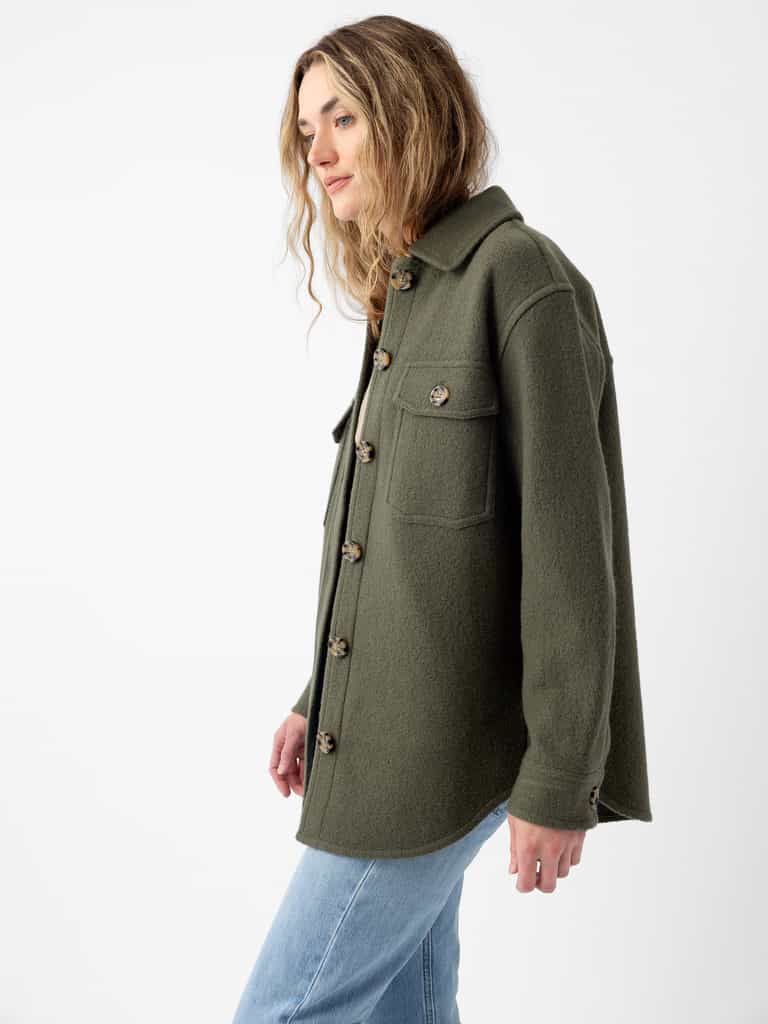 A person with long, wavy hair is wearing the Women's Boucle Shacket by Cozy Earth in a green color. The shacket features front pockets and a collar. They are standing in a three-quarter pose against a plain white background and are dressed in light blue jeans. |Color:Olive