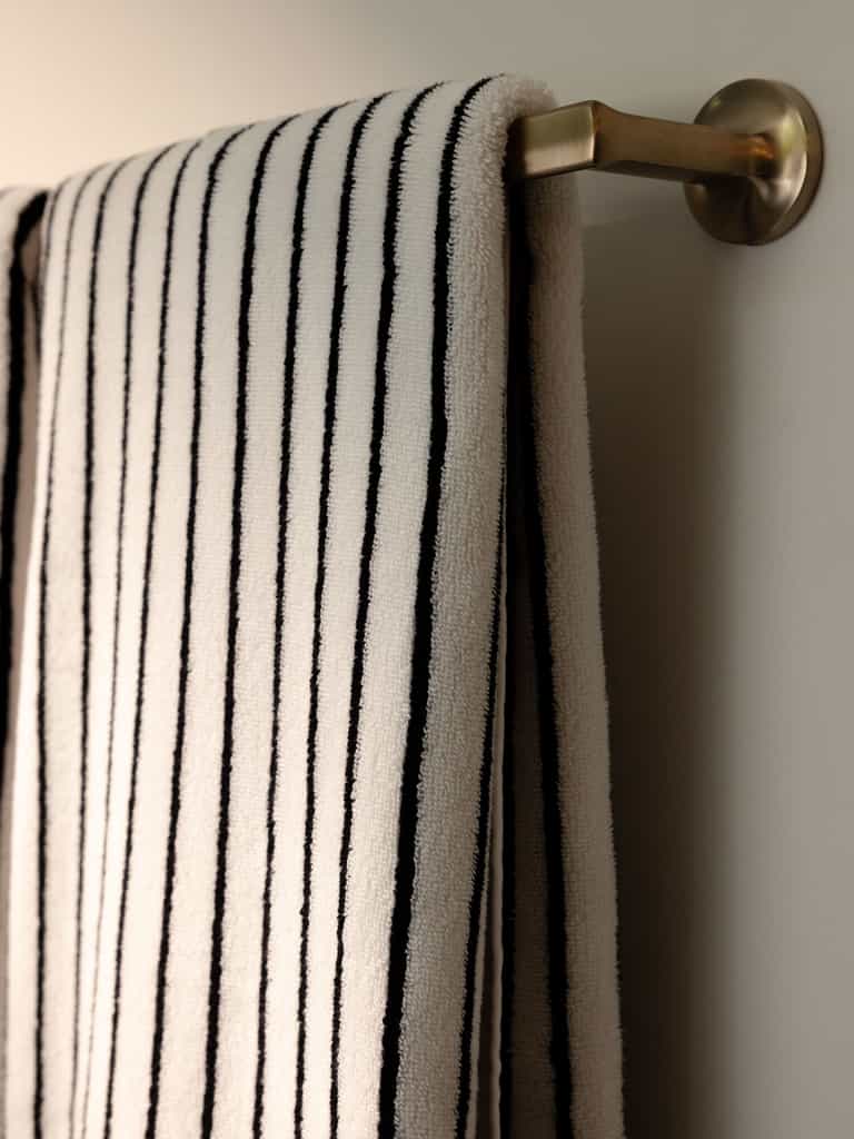 A Luxe Bath Towel from Cozy Earth, featuring elegant black vertical stripes on a pristine white fabric, hangs neatly on a brass towel rod mounted on a white wall. Soft lighting casts gentle shadows, accentuating the towel's plush texture. |Color:Black Stripe