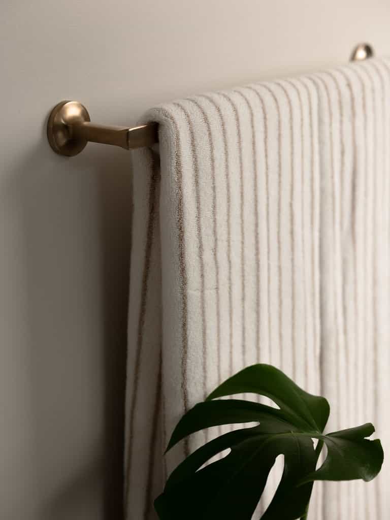A neatly folded Luxe Bath Towel in textured beige from Cozy Earth hangs on a brass towel bar mounted on a light-colored wall. Below the towel, a green Monstera plant leaf is partially visible, adding a touch of nature to the clean and minimal bathroom setting. |Color:Sand Stripe