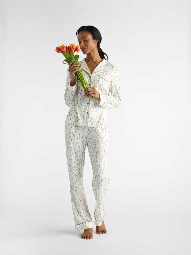 A woman in Cozy Earth Women's Bamboo Stretch-Knit Pajama Pant stands barefoot, inhaling the scent of orange tulips against a white backdrop. |Color:Sienna Tulips