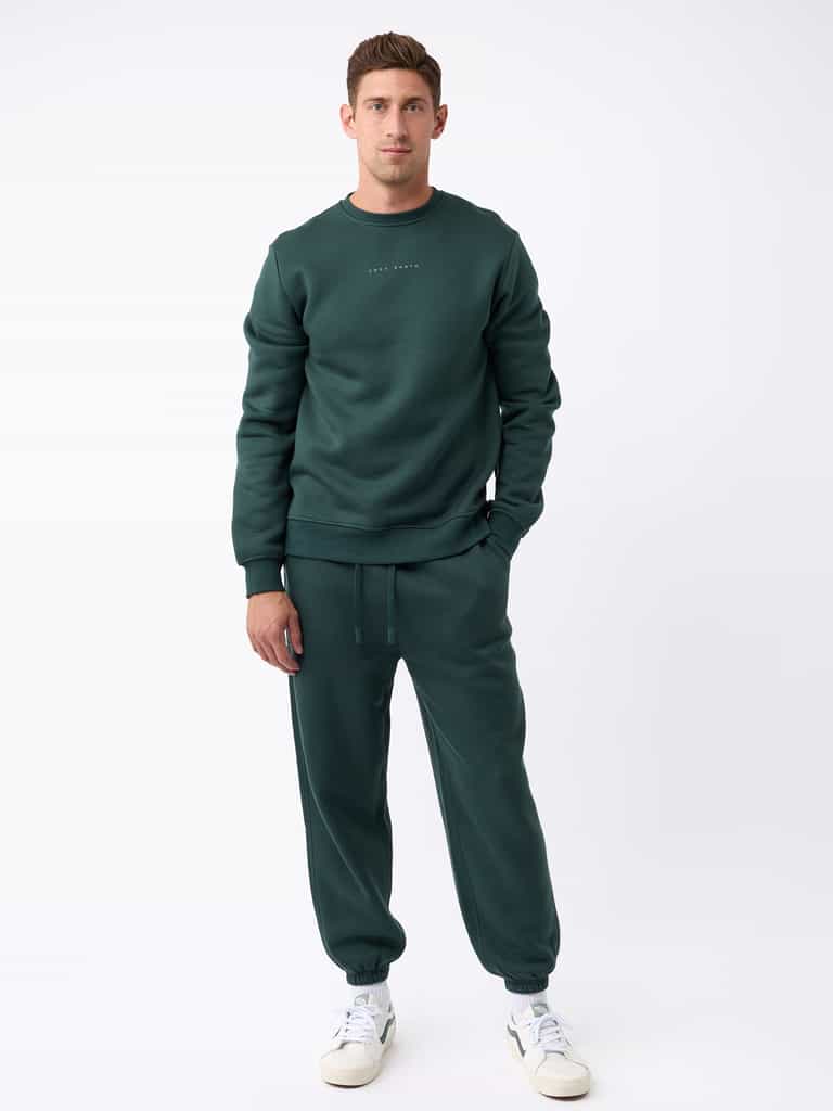 A man is standing against a white background wearing the dark green Men's CityScape Crewneck from Cozy Earth paired with matching joggers. He completes his look with white sneakers. |Color:Spruce