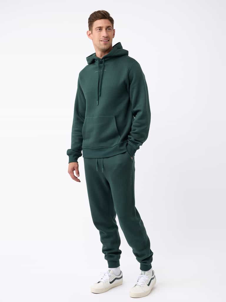 A person wearing the Men's CityScape Jogger by Cozy Earth in dark green stands against a plain white background. They are wearing white sneakers and have their hands in the jogger's hoodie pocket, looking slightly to the side. |Color:Spruce