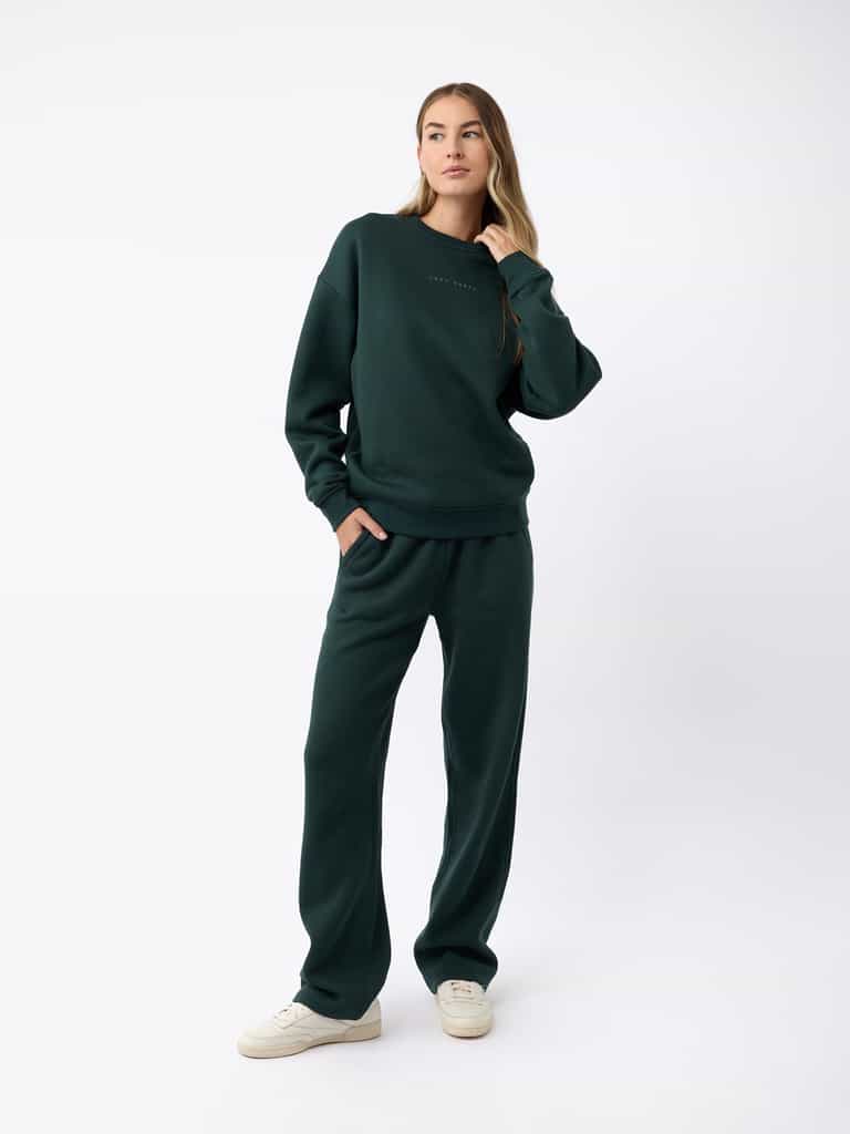 A person wearing the Women's CityScape Wide Leg Pant by Cozy Earth in a dark green color stands against a plain white background. They have long hair and are wearing white sneakers, with one hand in their pocket and a relaxed pose. |Color:Spruce