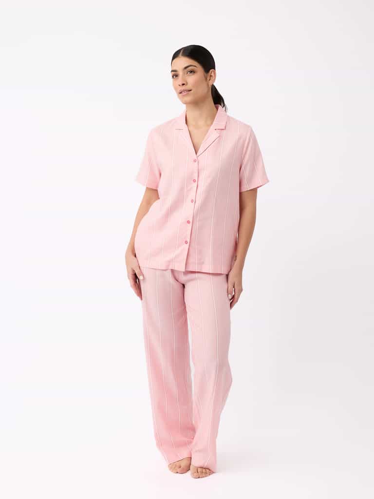 A person models a light pink, pinstriped pajama set from Cozy Earth. The short-sleeve top features buttons and the pants are relaxed fit, all posed against a white backdrop. |Color:Sweetheart Stripe