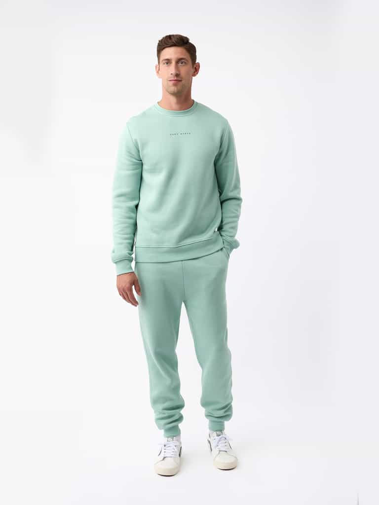 A person wearing the Cozy Earth's Men's CityScape Crewneck in light green, along with matching sweatpants, stands against a plain white background. They are also sporting white sneakers. The outfit looks casual and comfortable. |Color:Wintergreen