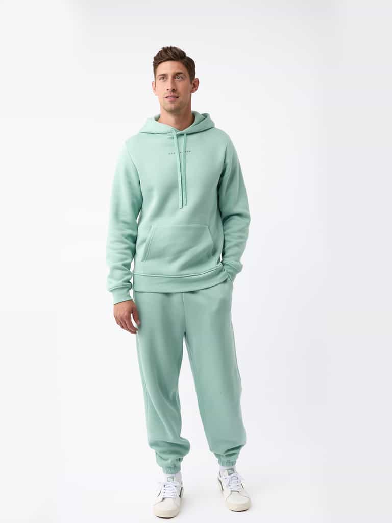 A person is wearing the Men's CityScape Hoodie from Cozy Earth in mint green, paired with matching sweatpants, standing against a plain white background. They complete their outfit with white sneakers featuring green details. |Color:Wintergreen
