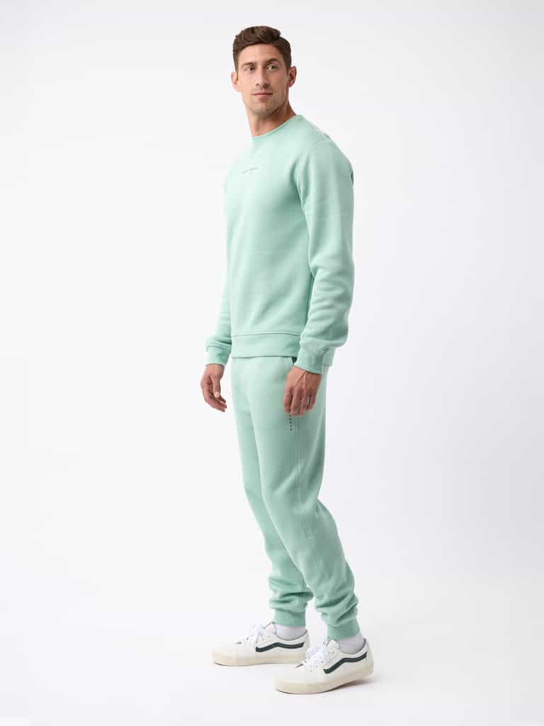 A person stands against a white background, dressed in the Men's CityScape Jogger from Cozy Earth, along with white sneakers. The individual looks at the camera with a neutral expression, and the jogger features minimal branding. |Color:Wintergreen