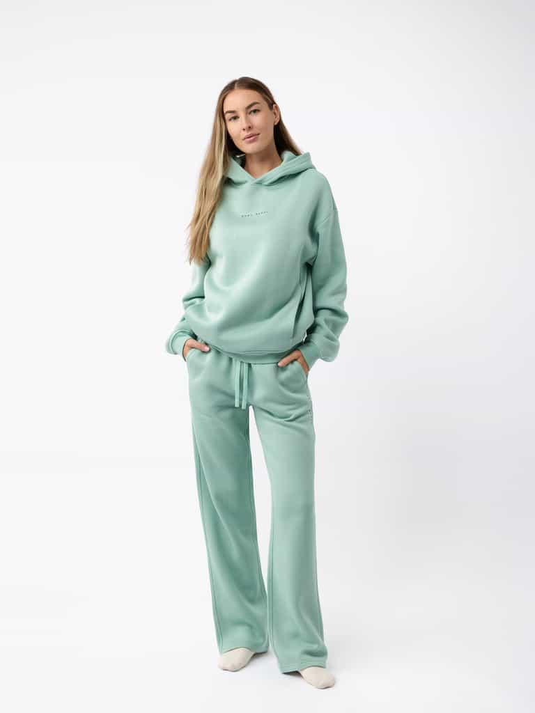 A person stands against a plain backdrop, wearing a coordinated light green hoodie paired with Cozy Earth's Women's CityScape Wide Leg Pant. Their hands rest in the hoodie pockets and their long, light brown hair cascades down, completing a casual and relaxed appearance. |Color:Wintergreen