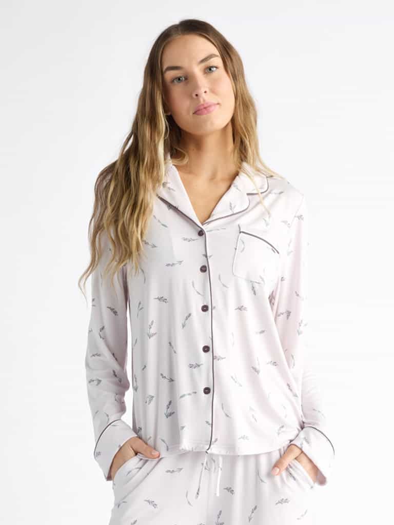 A person wearing Cozy Earth Women's Bamboo Stretch-Knit Long Sleeve Pajama Top, featuring a collar, buttons, and chest pocket, stands relaxed against a white background with long hair and hands in pockets. The light-colored top has a subtle print. |Color:French Lavender