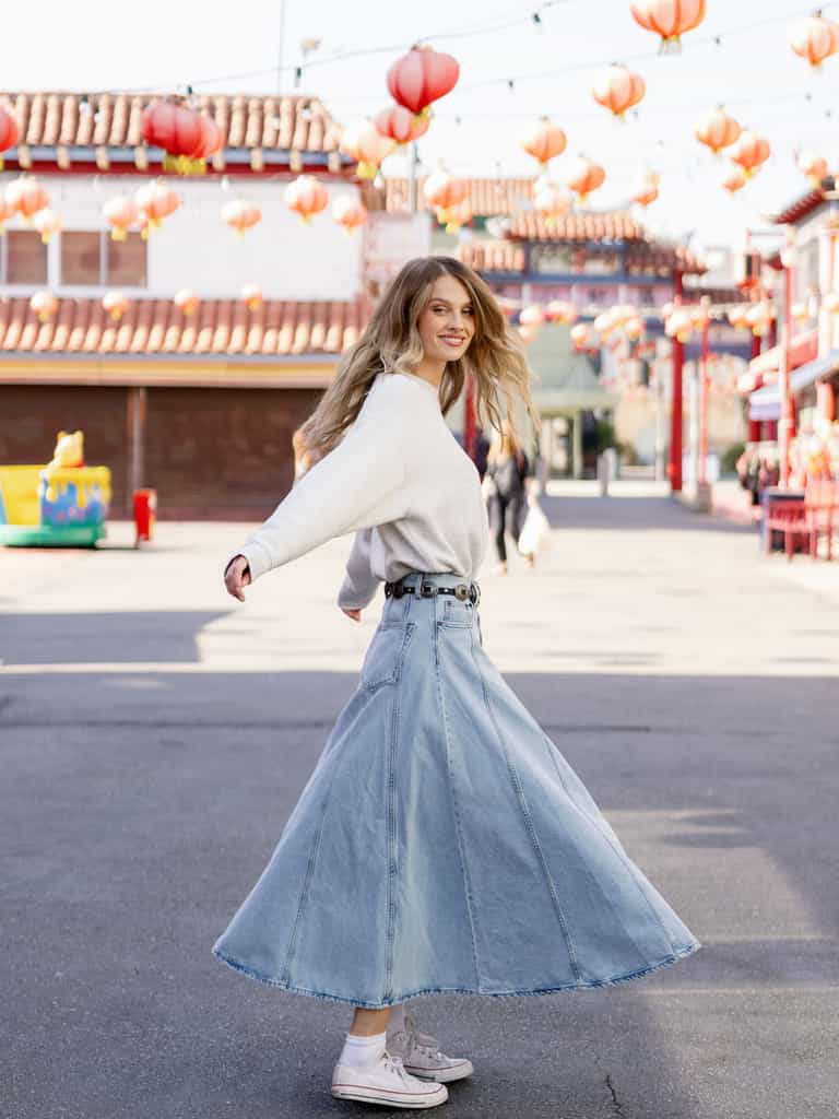 Heather Grey CityScape Pullover Crew. The Pullover is being worn by a female model. An accompanying jean skirt is being worn to complete the look of the outfit. The photo was taken with a downtown city background. |Color:Heather Grey