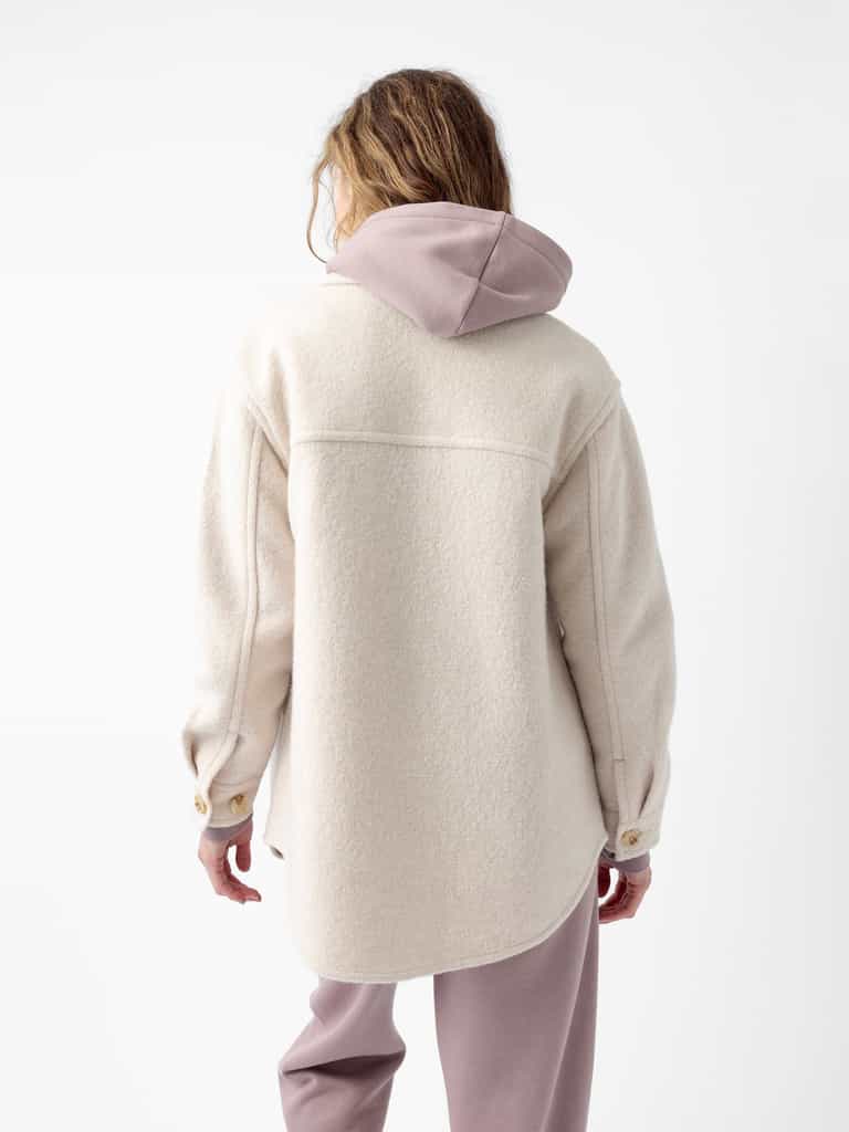 A person stands with their back to the camera, wearing the Cozy Earth Women's Boucle Shacket, an oversized light cream wool jacket featuring brown buttons and a pink hood. They are also dressed in matching pink pants, with a plain white backdrop in the background. |Color:Alabaster