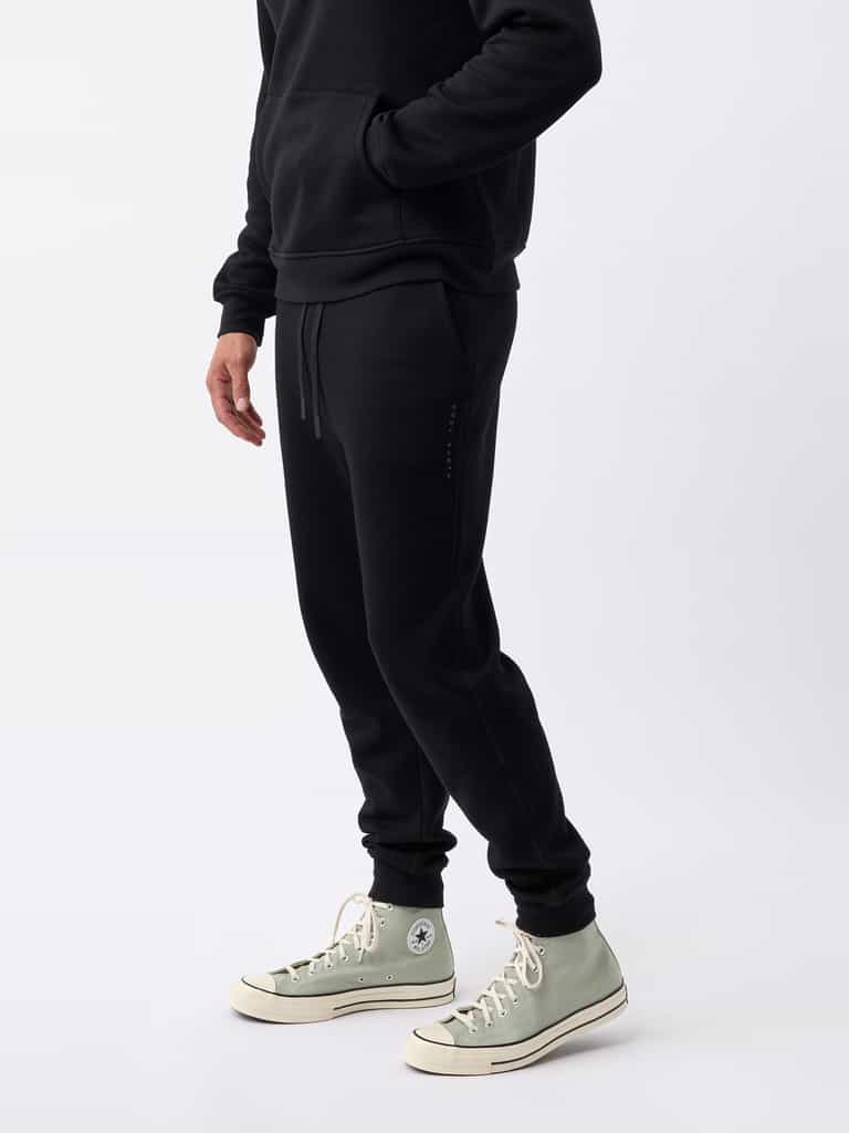 A person wearing Cozy Earth's Men's CityScape Jogger in black and a matching black hoodie stands against a plain white background. They are also sporting light green high-top sneakers, though their face is not visible in the image. |Color:Black