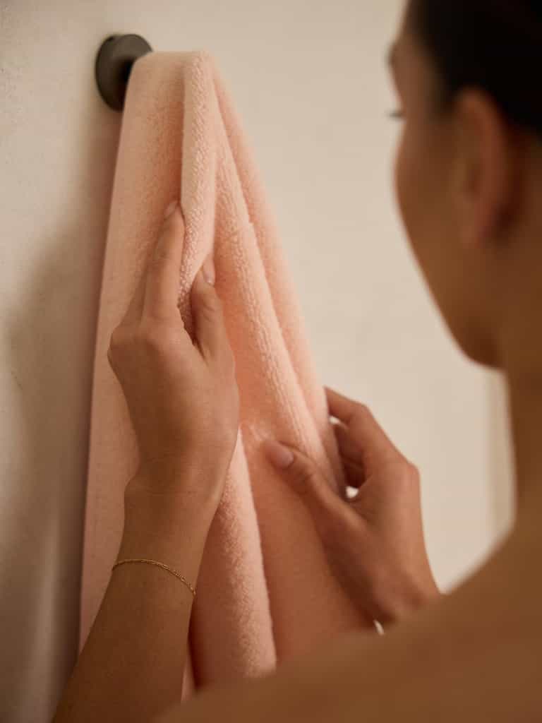 Wearing a delicate bracelet, a person hangs a Luxe Bath Towel by Cozy Earth on a wall hook. The focus is on the soft pink towel and their hands, set against a muted background. |Color:Blossom