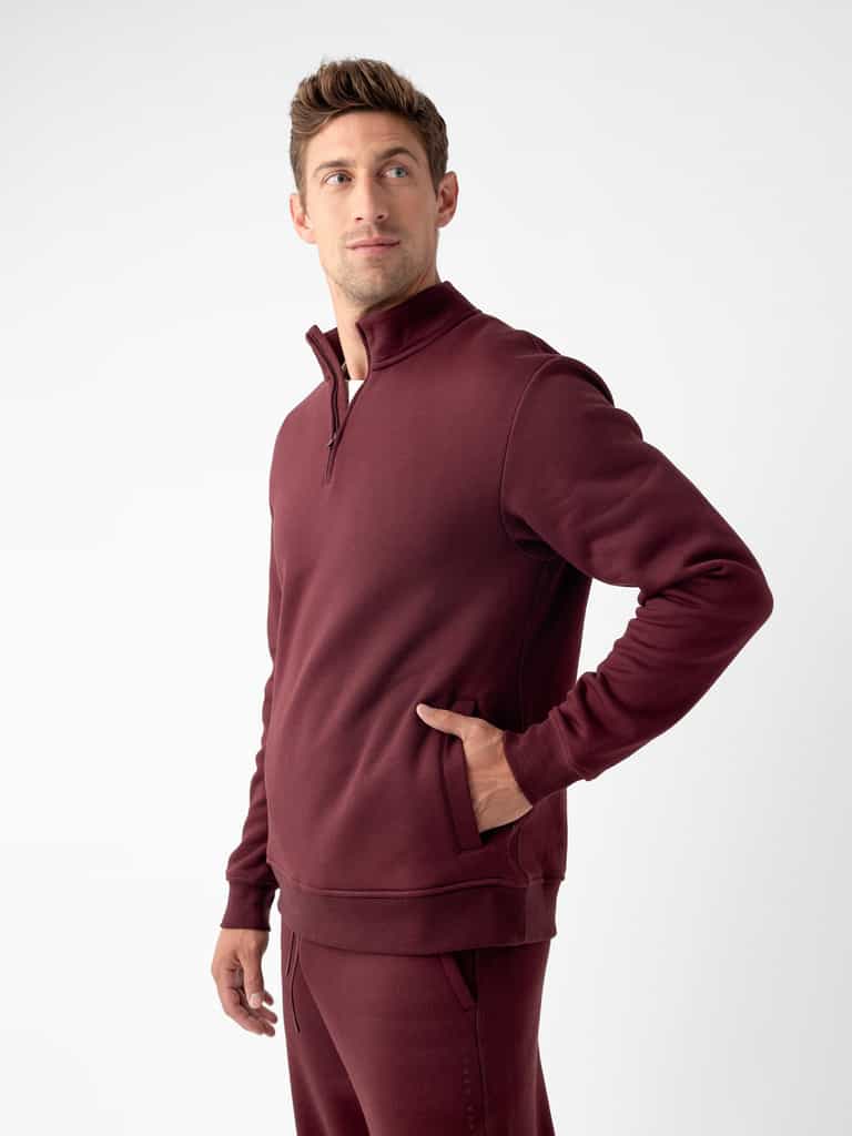 A person wearing a matching maroon Cozy Earth Men's CityScape Quarter Zip and pants is looking upwards and to the side. The individual has their right hand in the pocket of the sweatshirt. The background is plain and white. |Color:Burgundy