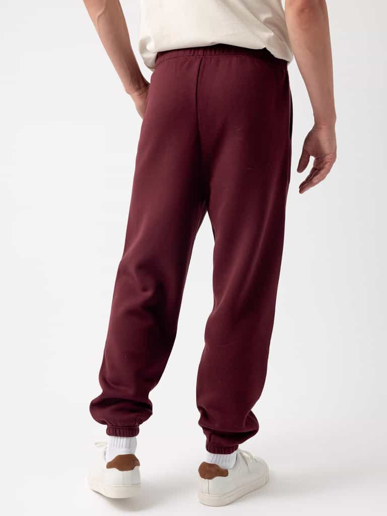 A person in a white shirt stands with their back to the camera, wearing the Cozy Earth Men's CityScape Sweatpant in maroon, which features elastic cuffs, paired with white sneakers that have brown accents. The background is plain white. |Color:Burgundy