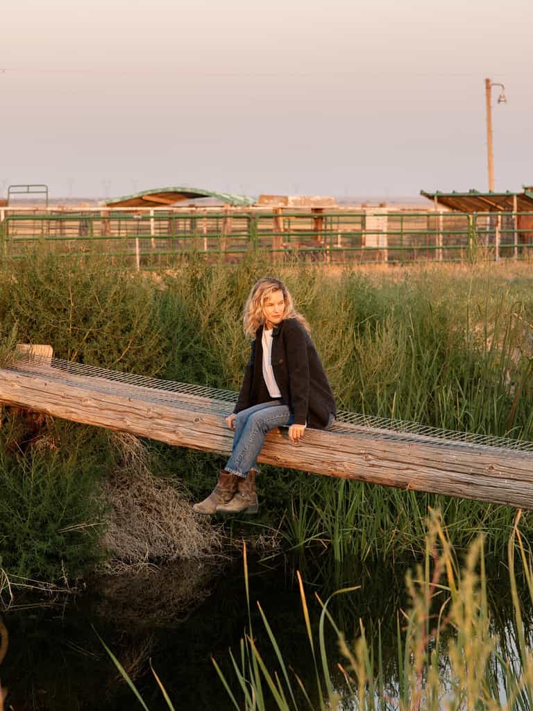 A person with short, wavy blonde hair sits on a wooden beam over a grassy and watery landscape. They wear a Cozy Earth Women's Boucle Shacket, a white shirt, blue jeans, and boots. In the background, there are green and brown agricultural structures under an evening sky. |Color:Coal