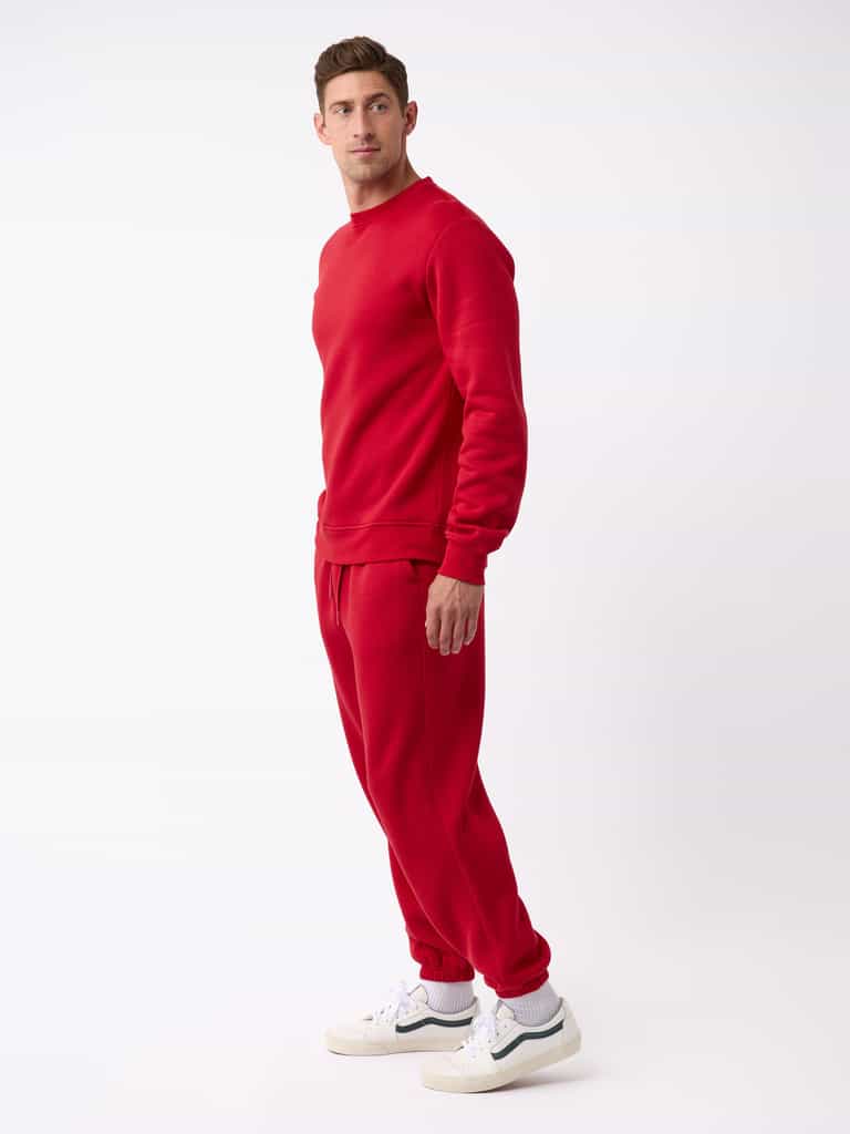 A man stands against a plain background wearing the Men's CityScape Sweatpant from Cozy Earth and a matching red sweatshirt. He is facing slightly to the side, with his right hand visible. He is also wearing white sneakers with black details. |Color:Crimson