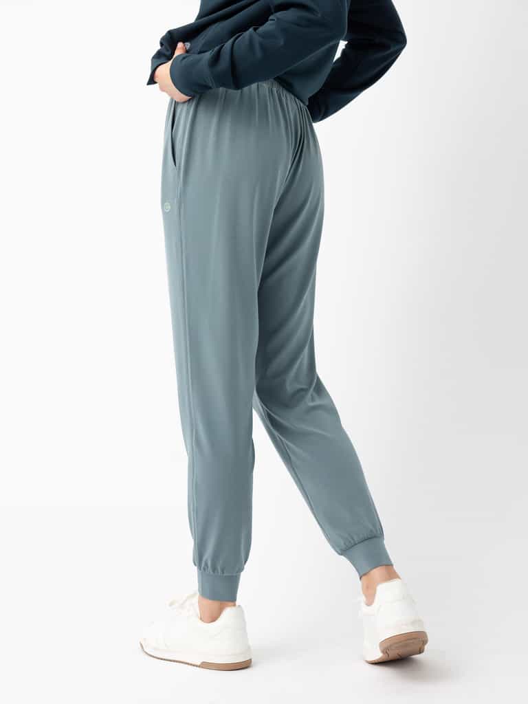 A person stands with one hand in their pocket, wearing Women's Studio Jogger pants by Cozy Earth in a teal color, a dark long-sleeve top, and white sneakers. The joggers feature a relaxed fit with an elastic waistband and cuffs at the ankles. The person is posing against a plain white background. |Color:Deep Blue