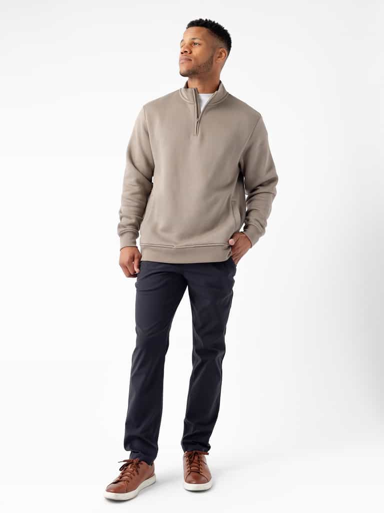 A man stands against a plain white background, gazing upward to his right. He is wearing Cozy Earth's Men's CityScape Quarter Zip in taupe over a white shirt, paired with black pants and brown leather sneakers. His hands are in his pockets, exuding a calm expression. |Color:Ebony