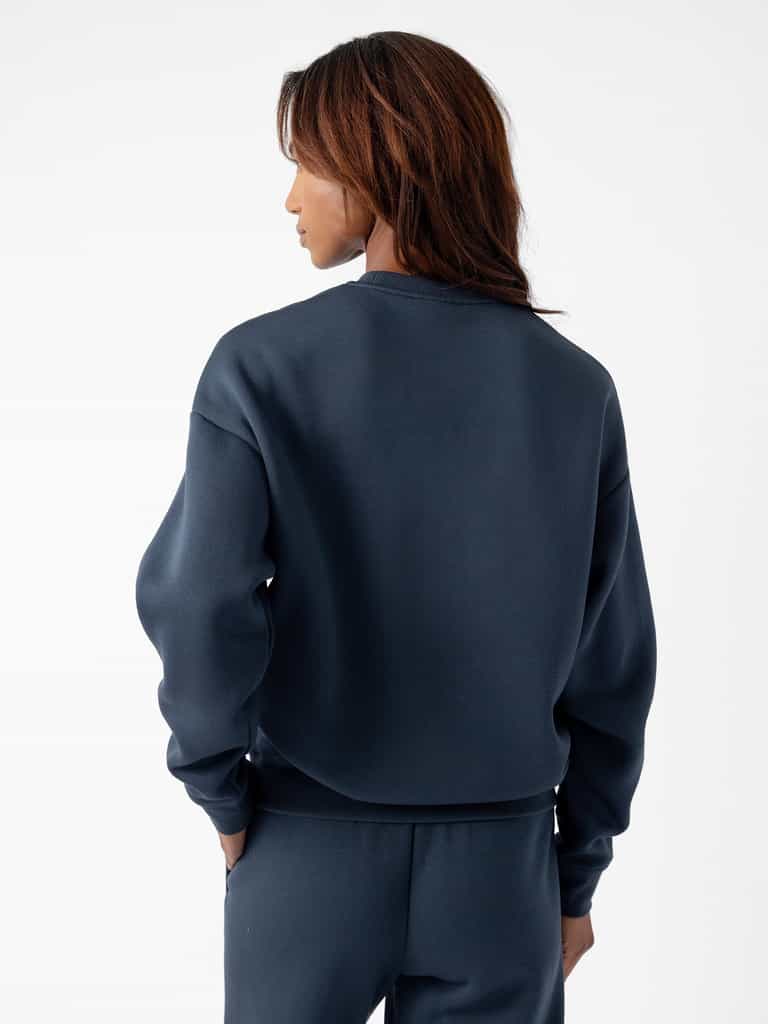 A person with shoulder-length, dark brown hair is seen from the back, wearing a dark blue Women's CityScape Crewneck by Cozy Earth and matching pants. They are standing against a plain white background. |Color:Eclipse