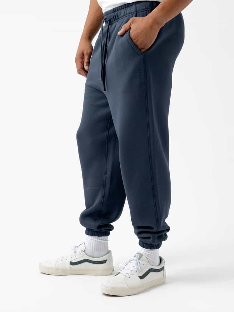 A person wearing Cozy Earth's Men's CityScape Sweatpant in dark blue with pockets, a white T-shirt, white socks, and white sneakers with dark accents stands against a plain white background. The person's hands are in the pockets of the sweatpants. |Color:Eclipse
