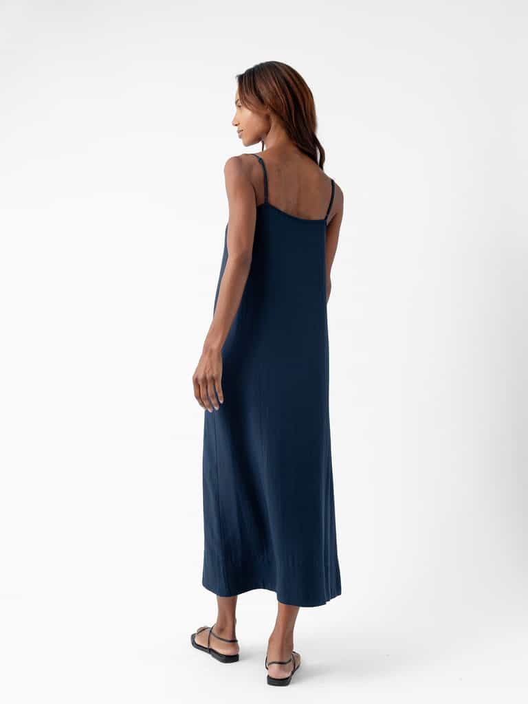 A woman with long hair is standing against a plain white background. She is wearing the Women's Sunset Slip Dress in navy blue from Cozy Earth and black flip-flops. The woman is turned slightly to the side, looking towards the right, showcasing the back of her dress. |Color:Eclipse