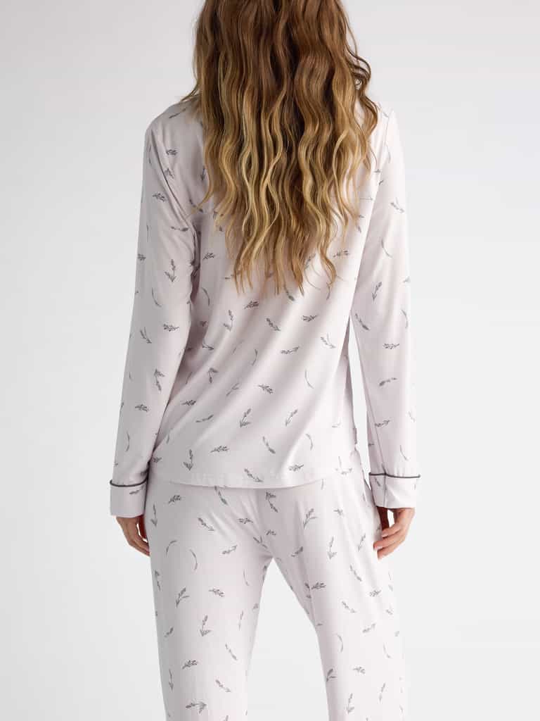 A person with long wavy hair faces away, wearing Cozy Earth Women's Bamboo Stretch-Knit Long Sleeve Pajama Top in a light color featuring a subtle pattern against a plain white background. |Color:French Lavender