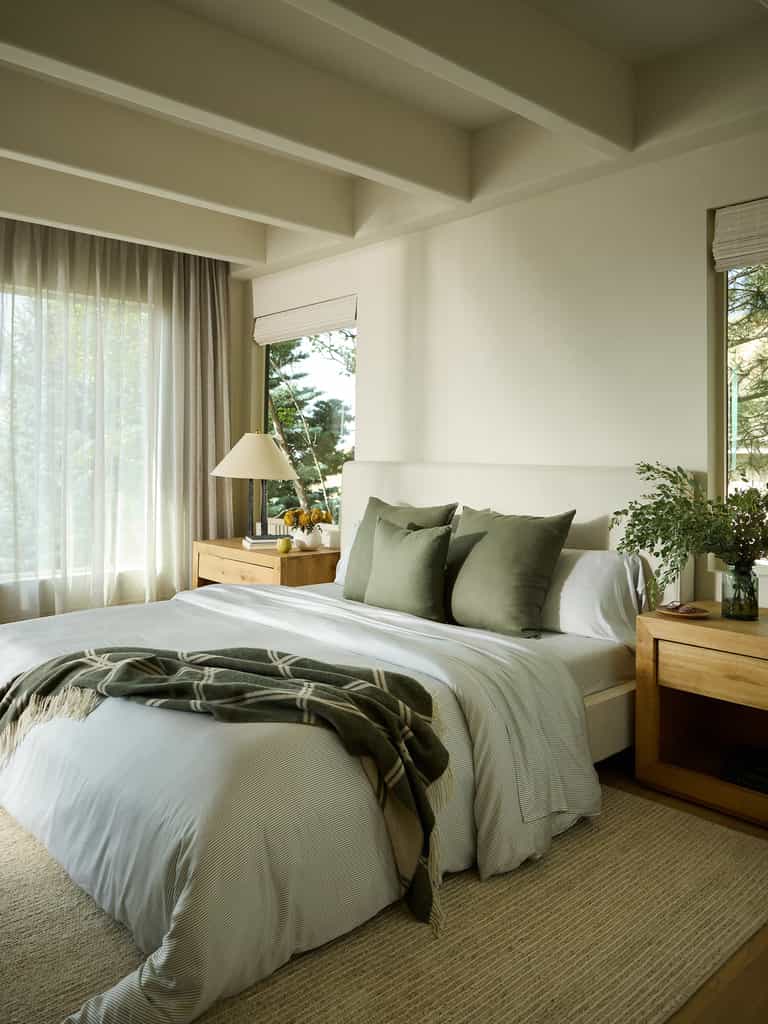 A cozy bedroom with a neatly made bed showcasing white Bamboo Flat Sheets from Cozy Earth and green pillows. A green throw is draped over the bed. Wooden nightstands hold a lamp and a vase with greenery, while sunlight filters through sheer curtains next to large windows. |Color:Jade Pinstripe