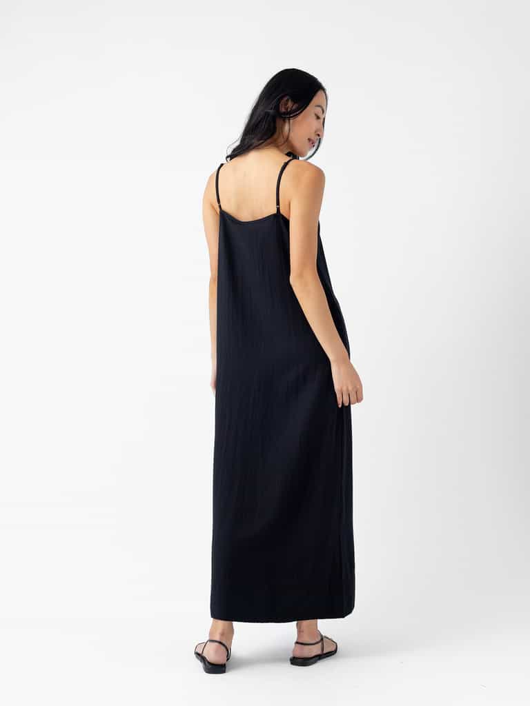 A woman with long black hair wearing the Cozy Earth Women's Sunset Slip Dress and black sandals stands facing away, looking over her right shoulder against a plain white background. |Color:Jet Black