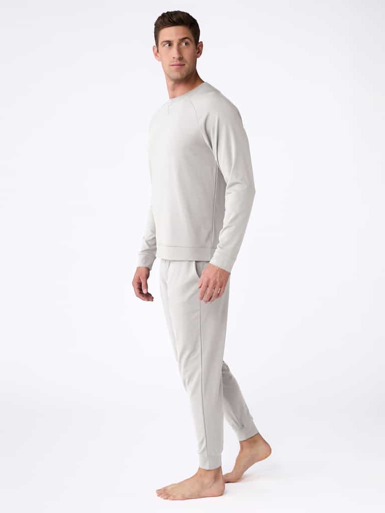 Modeling on a plain white background, an individual is barefoot, dressed in the Men's Bamboo Jogger Set from Cozy Earth. They wear a light gray long-sleeve sweatshirt and matching sweatpants while turned slightly to the side, gazing forward. |Color:Light Grey
