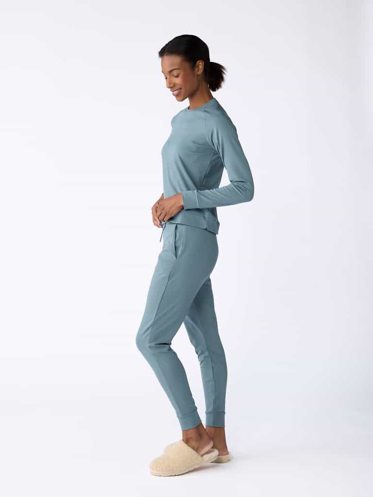 A woman wearing Cozy Earth's Women's Bamboo Jogger Pant in teal and beige slippers smiles while standing in a relaxed pose against a white background. Her hair is pulled back as she stands in profile, with her hands resting on her hips. |Color:Oceanside