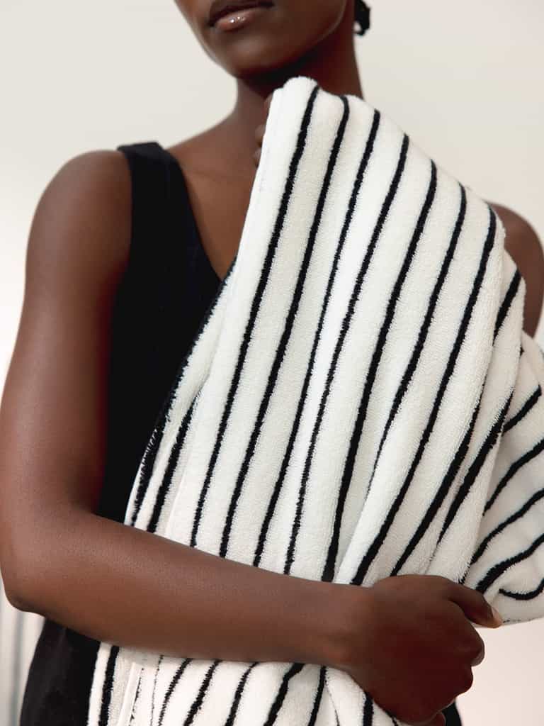 A person wearing a black sleeveless top holds a Luxe Bath Towel by Cozy Earth, featuring black stripes on white fabric, draped over one shoulder. The background is blurred and neutral. The person's face is partially shown from the nose down. |Color:Black Stripe