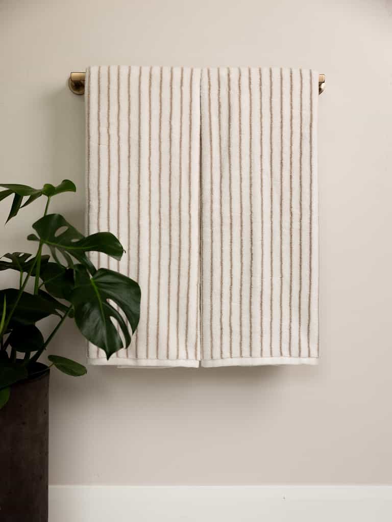 A Cozy Earth Luxe Bath Towel featuring thin, vertical beige stripes hangs neatly on a brass towel bar against a light, neutral-colored wall. To the left of the towel, a green potted plant with large leaves adds a natural touch. The overall scene is minimalist and calming. |Color:Sand Stripe