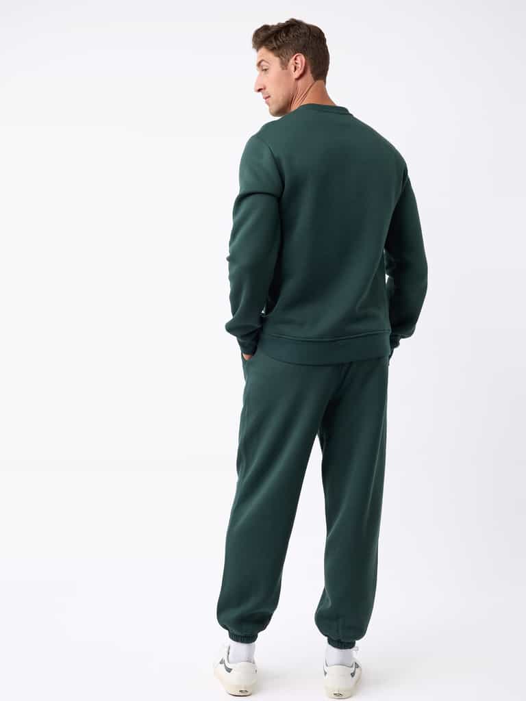 A man, with short brown hair, stands facing slightly away from the camera in a white studio setting while wearing the Men's CityScape Crewneck and matching sweatpants from Cozy Earth in dark green. |Color:Spruce