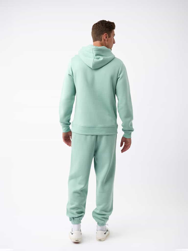A person wearing a Men's CityScape Hoodie in light green from Cozy Earth, with matching sweatpants, stands facing away from the camera on a plain white background and is sporting white sneakers. |Color:Wintergreen