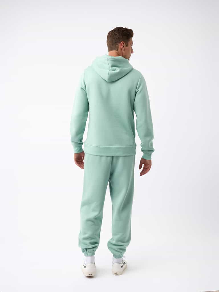 A person dressed in a Men's CityScape Sweatpant from Cozy Earth is facing away from the camera. They are standing against a plain white background, showcasing their light green hoodie and matching sweatpants, along with visible white sneakers. |Color:Wintergreen
