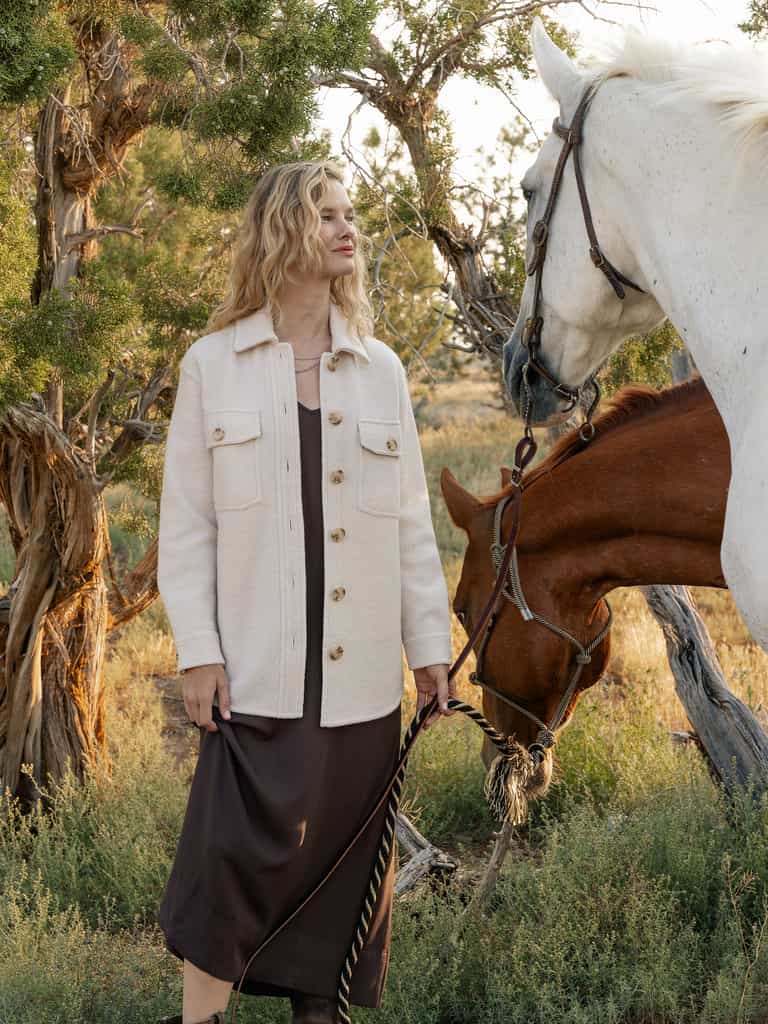 A person with wavy blonde hair stands outdoors wearing a Cozy Earth's Women's Boucle Shacket in beige over a brown dress, holding the reins of two horses. The background features trees and grass, suggesting a natural setting. |Color:Alabaster