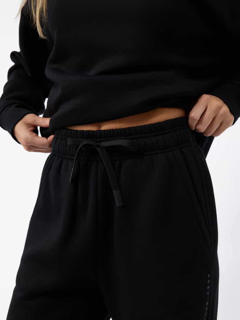 A person wearing a black Women's CityScape Wide Leg Pant from Cozy Earth adjusts the waistband. The photo focuses on their midsection, highlighting the drawstring and fabric texture against a plain white background. |Color:Black