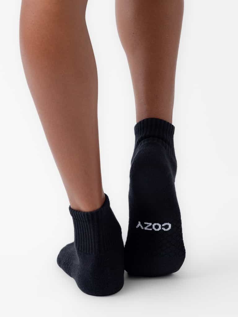 A person wearing black Essential Quarter Socks by Cozy Earth stands on a white surface. The word "COZY" is printed in white on the sole of the right sock. |Color:Black