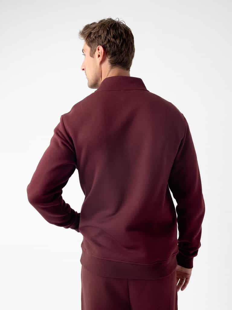 A person with short hair is standing with their back to the camera, wearing a maroon Men's CityScape Quarter Zip and matching pants by Cozy Earth. The plain white background highlights the outfit. They have one hand on their hip. |Color:Burgundy