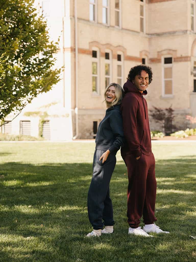 Two people stand back-to-back on a grassy lawn, both smiling. One is wearing a navy blue hoodie and a pair of Cozy Earth's Men's CityScape Sweatpants, while the other is dressed in a maroon hoodie and matching Men's CityScape Sweatpants from Cozy Earth. A tall, light-colored building and a tree can be seen in the background. |Color:Burgundy