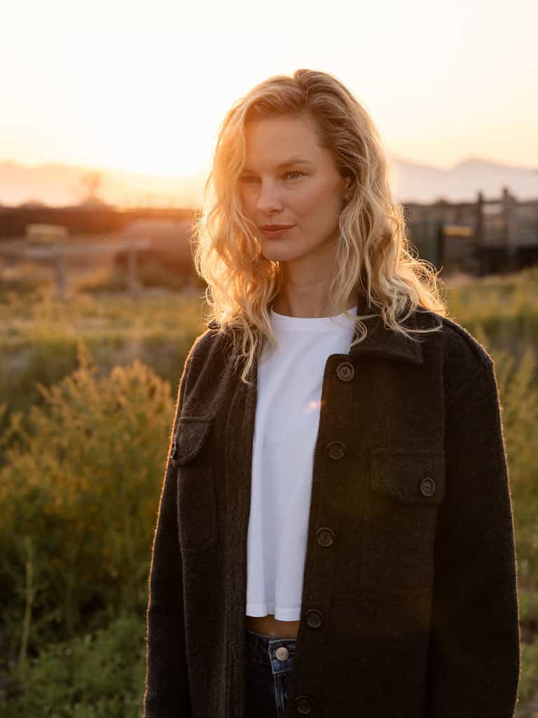 A woman with wavy blonde hair stands outdoors at sunset, wearing a dark coat over a white shirt. She is looking slightly to the side. The background features fields, some plants, and distant mountains bathed in the warm light of the setting sun. She is elegantly dressed in a Cozy Earth Women's Boucle Shacket. |Color:Coal