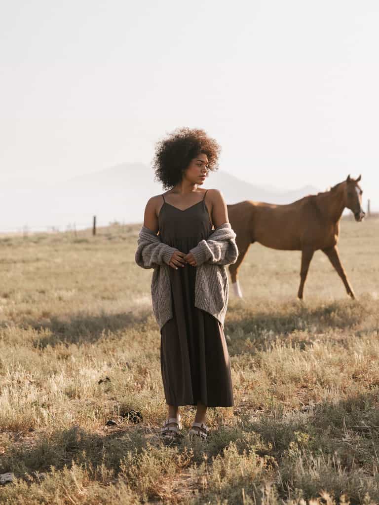 A person with curly hair stands in a grassy field wearing Cozy Earth's Women's Sunset Gathered Dress and a light gray cardigan, with eyes gazing downward. In the background, a brown horse walks casually. Misty mountains are visible in the distance under a pale sky. |Color:Cacao