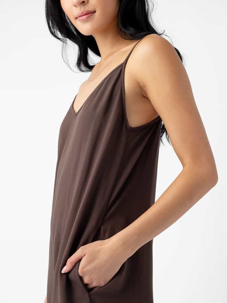 A person with long dark hair wears a Cozy Earth Women's Sunset Slip Dress, featuring a sleeveless design and dark brown color with a V-neckline. Their right hand is casually placed in the dress pocket, and they are slightly turned to one side against a plain white background. |Color:Cacao
