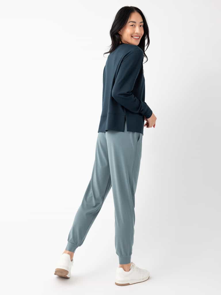 A person with long, dark hair smiles while posing in a dark green jacket and Cozy Earth's Women's Studio Jogger in light blue. They are standing against a plain white background and wearing white sneakers. Their body is turned slightly to the side, and they are looking back at the camera. |Color:Deep Blue