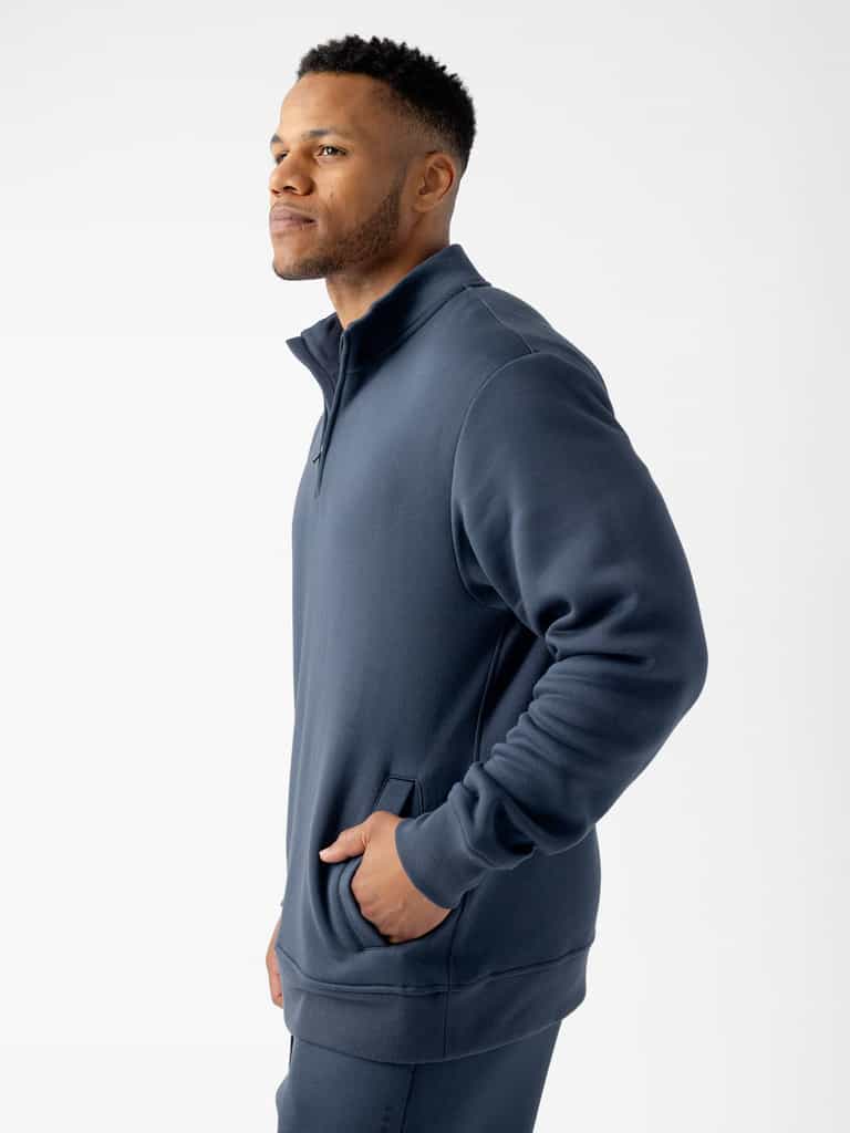 A man with short hair is standing against a plain white background, wearing a Cozy Earth Men's CityScape Quarter Zip in navy blue. He has his left hand in his pocket and is looking to the side. |Color:Eclipse