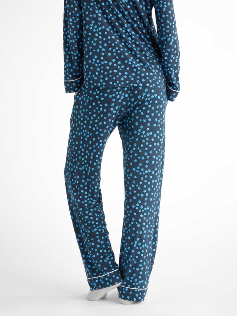 A person is wearing the Women's Bamboo Stretch-Knit Pajama Pant by Cozy Earth, featuring a dark blue background with a pattern of small blue flowers. The image captures their back view from the neck down, standing on a neutral-colored surface. |Color:Forget Me Nots