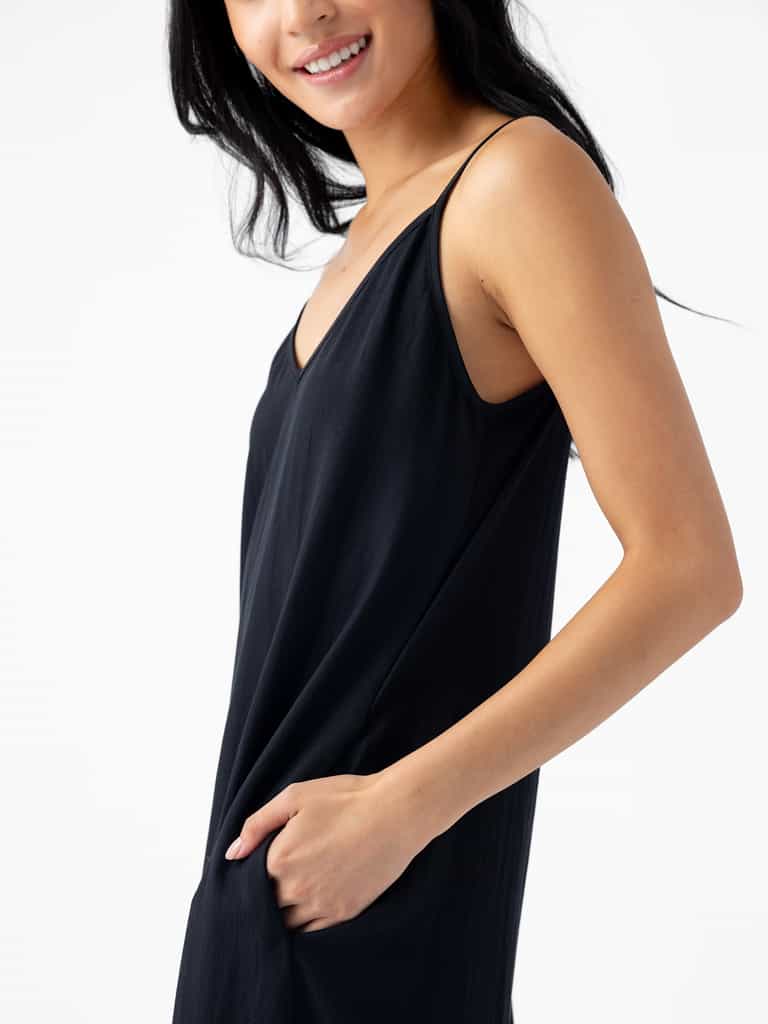A person with long black hair smiles and wears the Women's Sunset Slip Dress by Cozy Earth. The person has one hand in a pocket, and the background is white. |Color:Jet Black