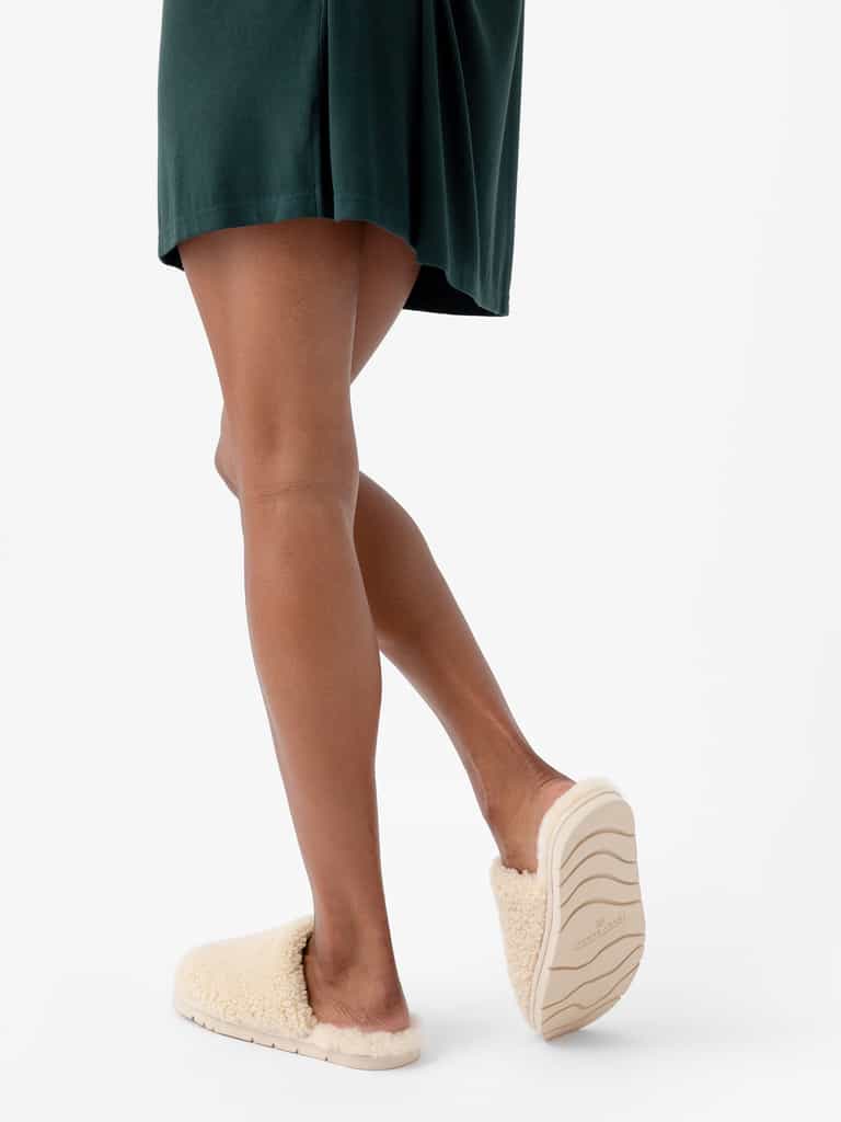 A person wearing a dark green dress is shown walking away from the camera. They are wearing a pair of Cozy Earth's Puffy Sheep Slippers, which are fluffy and cream-colored with light-colored soles. The white background emphasizes the attire and footwear. |Color:Natural